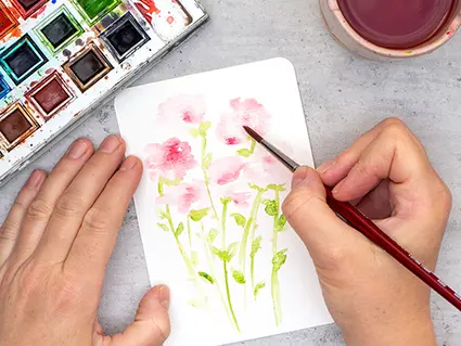 Jemand malt mit Aquarellfarben rote Rosen auf eine Aquarellpostkarte von Hahnemühle. Ebenfalls im Bild zu sehen ist ein Aquarellfarbkasten und ein Wasserbehälter aus Keramik.
