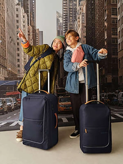 Ein Junge und ein Mädchen im Teenageralter stehen mit ihren Reisetrolleys von satch vor einer New-York-Kulisse. Das Mädchen zeigt mit dem Finger nach oben.