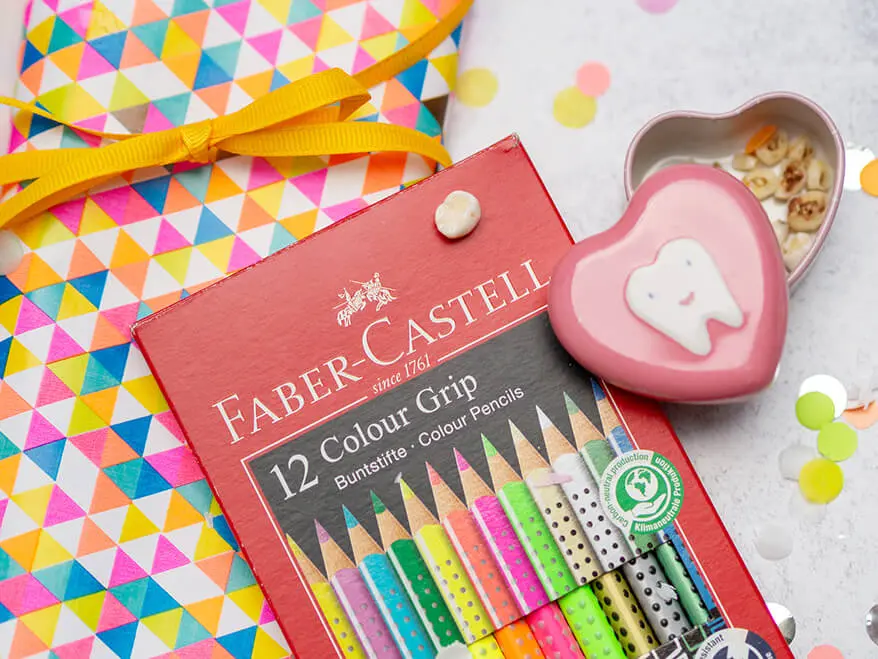 Ein Packung Buntstifte von Faber-Castell liegt auf einem bunten Geschenkpapier. Daneben steht eine rosafarbene Zahndose. Foto: Johanna Rundel