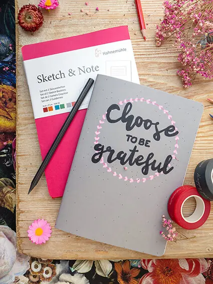 Auf einem rustikalen Holzbrett liegen zwei Notizbücher „Sketch & Note“ von Hahnemühle. Das Cover eines Buches wurde belettert.