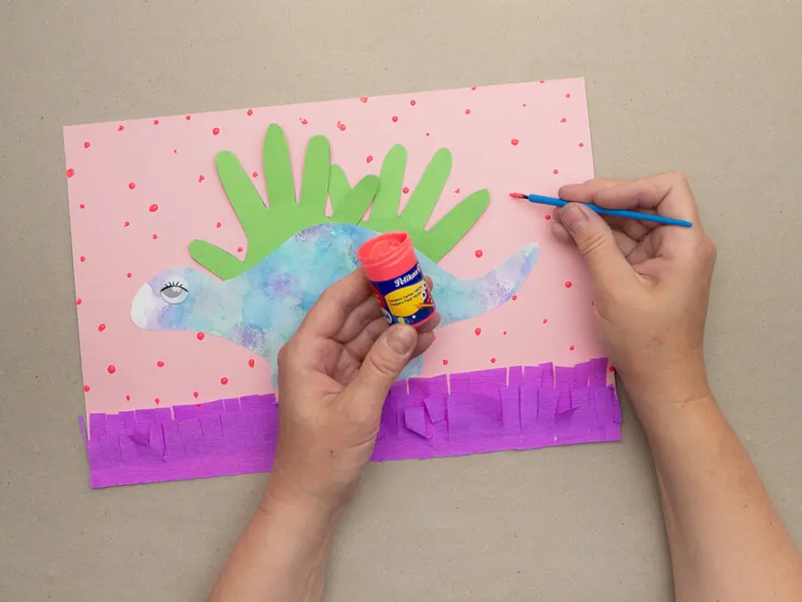 Jemand malt mit neonpinker Temperafarbe von Pelikan kleine Punkte auf den Hintergrund eines Dinosaurierbilds aus Handabdrücken. Foto: Johanna Rundel 