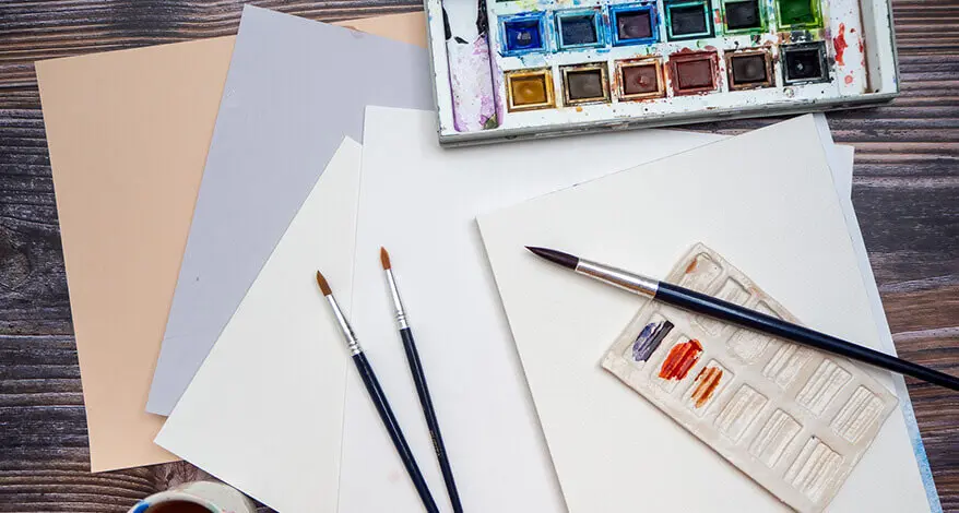 Auf einem dunklen Holztisch sind verschiedene Aquarellpapiere von Hahnemühle ausgebreitet. Pinsel und Aquarellfarbkasten liegen dabei. Foto: Johanna Rundel 