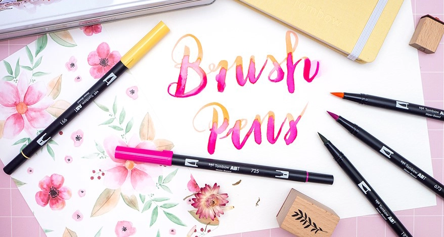 Auf einem Blatt Aquarellpapier wurde mit bunten Brush Pens von Tombow die Worte „Brush Pen“ aquarelliert. Verschiedene Stifte und Utensilien liegen darum verteilt. Foto: Johanna Rundel