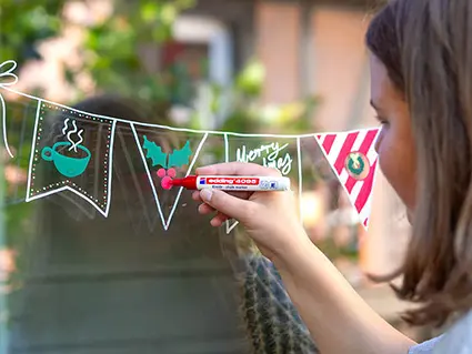 Eine junge Frau malt mit einem roten Kreidemarker von edding eine weihnachtliche Wimpelkette auf eine Fensterscheibe. Foto: Johanna Rundel