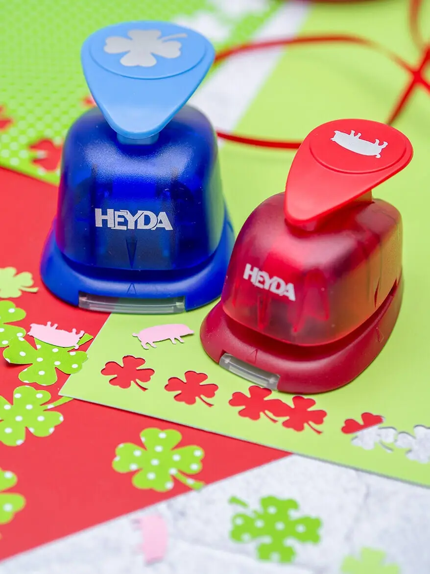 Ein blauer Kleeblatt- und ein roter Schweinchen-Stanzer von HEYDA stehen auf rotem und grünem Bastelpapier. Foto: Johanna Rundel