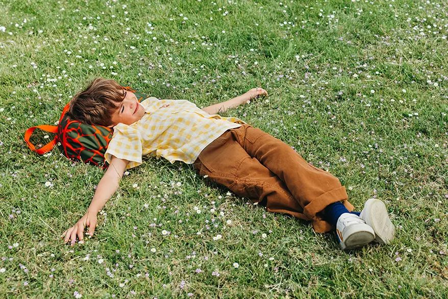 Ein Junge liegt ausgestreckt auf einer grünen Wiese mit dem Kopf auf seiner Sporttasche von ergobag.