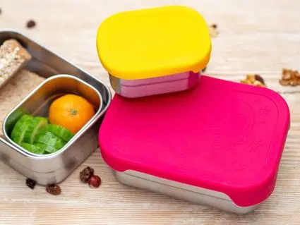 Drei Edelstahlbrotdosen von ergobag mit verschiedenfarbigen Silikondeckeln. Eine Dose ist geöffnet, man sieht einen Pausensnack aus Broten, Gurke und Mandarine. Foto: Johanna Rundel