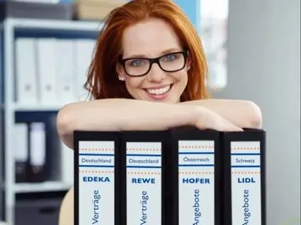 Eine rothaarige Frau mit Brille stützt sich mit ihren Unterarmen auf eine Reihe schwarzer Aktenordner mit gestalteten Etiketten von Avery Zweckform.