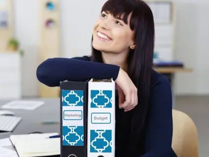 Eine junge, dunkelhaarige Frau stützt sich mit ihrem rechten Unterarm auf zwei schwarze Aktenordner mit gestalteten Etiketten von Avery Zweckform.