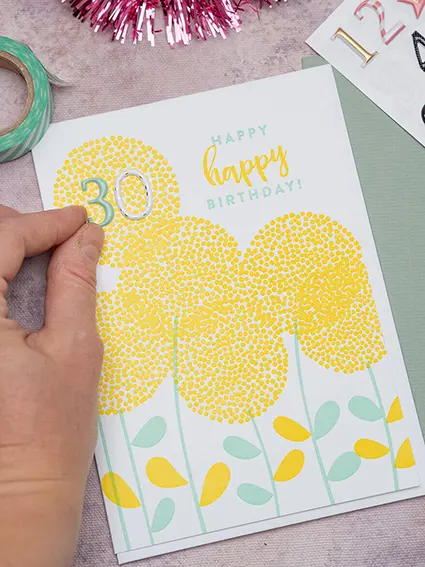 Jemand klebt die Zahlenaufkleber 3 und 0 auf eine Letterpress-Glückwunschkarte von Rössler mit gelben Blumen. Foto: Johanna Rundel 