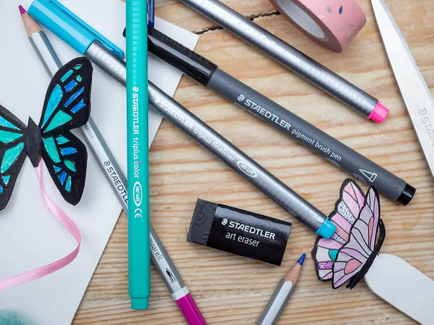 Auf einem Holzuntergrund liegen verschiedene Bunt- und Filzstifte der Marke STAEDTLER. Sie wurden zum Anmalen von zwei Lesezeichen in Schmetterlingsform verwendet. Foto: Johanna Rundel