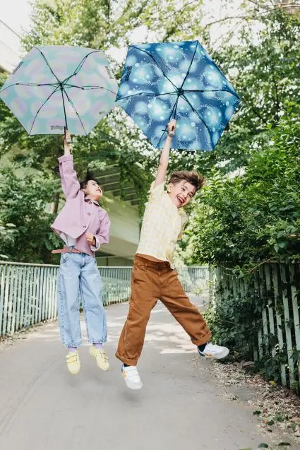 Zwei Kinder springen mit aufgespannten Regenschirmen der Marke ergobag in der Hand in die Luft.