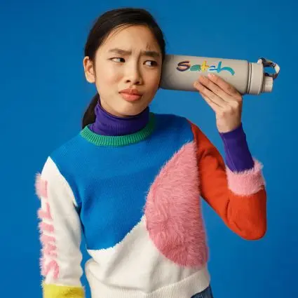 Eine Jugendliche in kunterbuntem Strickpulli hält sich eine Trinkflasche von satch ans Ohr und schaut skeptisch.