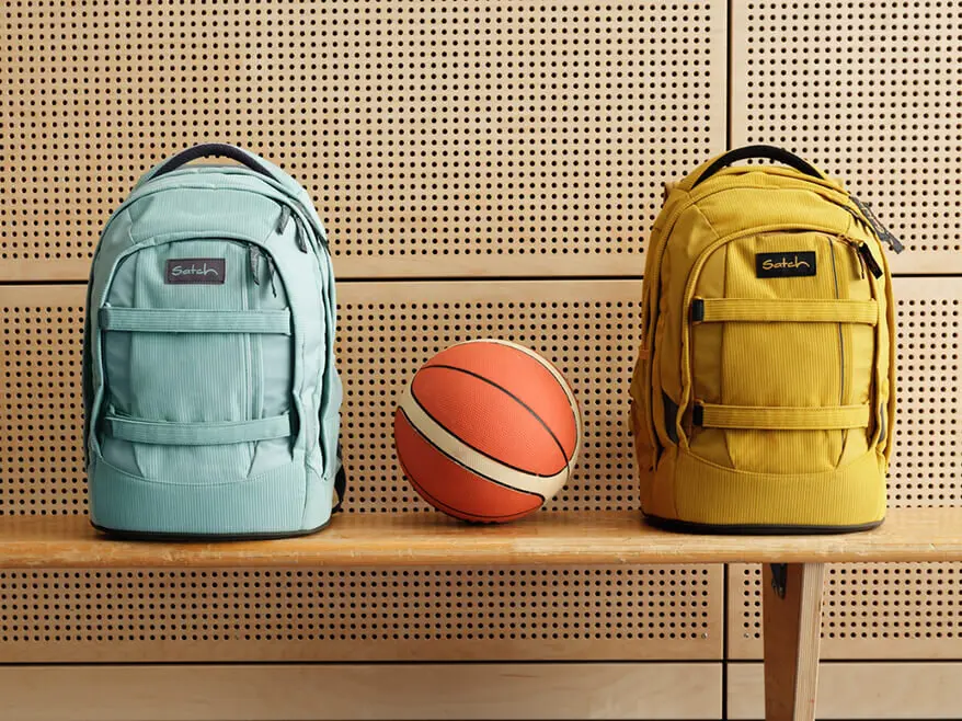 Zwischen eine hellblauen und einem gelben Schulrucksack von satch liegt ein Basketball auf einer Bank in einer Turnhalle.