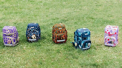 Fünf Schulranzen der Marke ergobag stehen nebeneinander aufgereiht auf einer grünen Wiese.