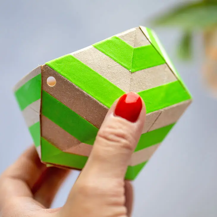 Eine Hand mit roten Fingernägeln hält einen selbstgebastelten, mit Washitape von HEYDA dekorierten Blumentopf aus einer Milchtüte hoch.