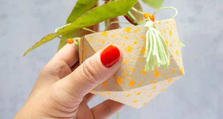 Eine Hand mit roten Fingernägeln hält einen selbstgebastelten, mit Washitape von HEYDA dekorierten Blumentopf aus einer Milchtüte hoch.