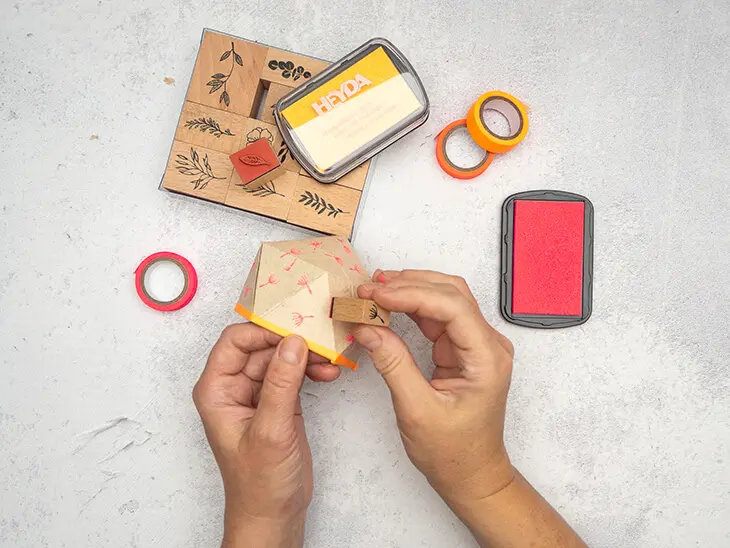 Jemand stempelt mit einem Stempel von HEYDA kleine Blümchen auf die Außenseite eines DIY-Blumentopfes aus einer Milchtüte.