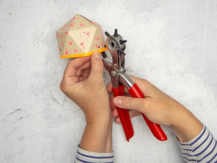 Jemand stanzt mit einer roten Lochzange von HEYDA kleine Löcher in einen selbstgebastelten Blumentopf aus einer Milchtüte.