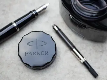 Ein geöffnetes Tintenfass, ein schwarzer Füller und ein Konverter liegen auf einer hellgrauen Filzmatte. In der Mitte liegt der Deckel des Tintenfasses mit der Aufschrift Parker. Foto: Johanna Rundel