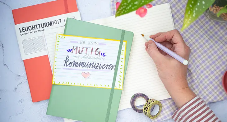 Jemand schreibt mit einem Drehgriffel von LEUCHTTURM1917 in ein Notizbuch. Weitere Notizbücher und eine Affirmationskarte liegen daneben. Foto: Johanna Rundel