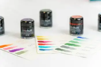 Auf einem hellen Untergrund stehen verschiedenfarbige Tintenfässer von Otto Hutt. Drei Musterkarten mit Farbstrichen liegen dazwischen.