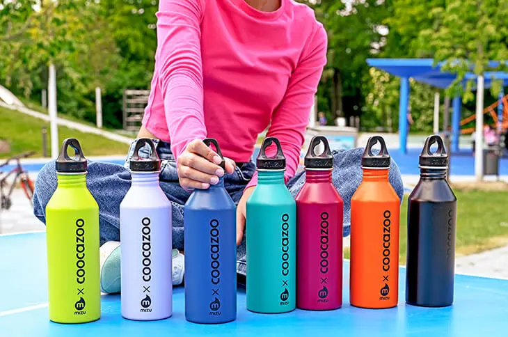 Eine Person im pinken Longsleeve greift eine Trinkflasche von coocazoo aus einer Reihe verschiedenfarbiger Trinkflaschen auf einer Tischtennisplatte.