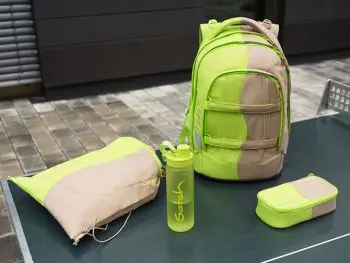 Schulrucksack, Sportbeutel, Trinkflasche und Mäppchen von satch mit beigen-neongelbem Design sind auf einer dunkelgrünen Tischplatte drapiert.