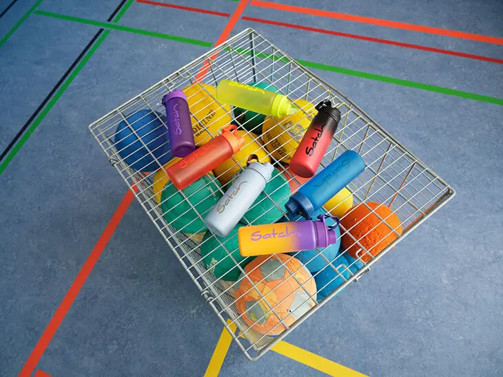 In einer Sporthalle steht ein Bällewagen aus Metall, darauf liegen viele verschiedene Trinkflaschen der Marke satch.