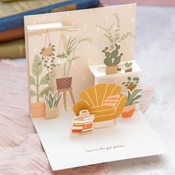 Eine quadratische Pop-Up-Karte von Grafik Werkstatt mit einem gemütlichen Sessel, vielen Büchern und Pflanzen sowie dem Satz „Lass es dir gut gehen“ steht aufgeklappt auf einem Tisch. Foto: Johanna Rundel