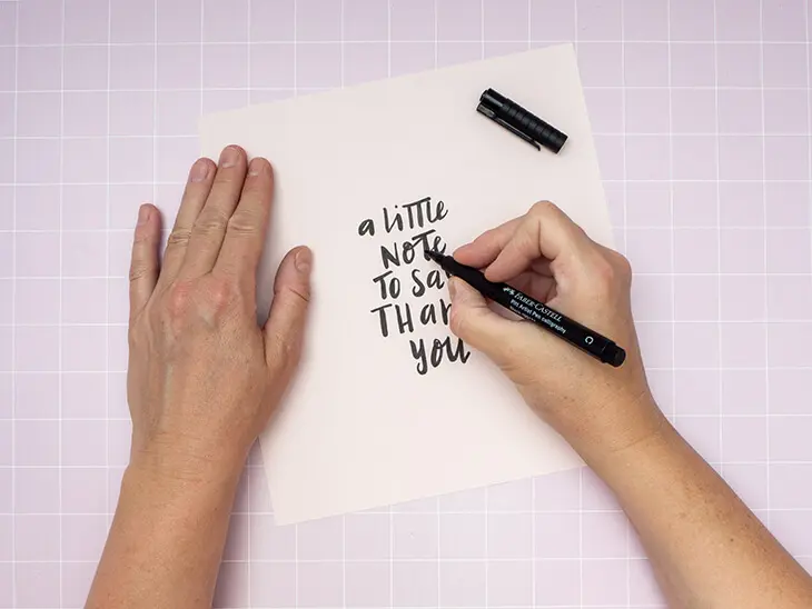 Jemand lettert mit einem schwarzen Kreativmarker von Faber-Castell den Satz „a little note to say thank you“ auf ein quadratisches Stück rosa Fotokarton. Foto: Johanna Rundel
