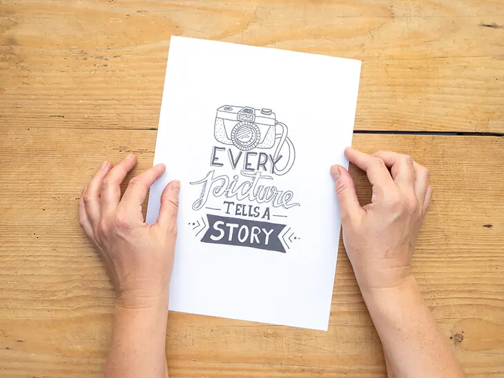 Jemand legt ein Blatt Papier mit dem Lettering „Every picture tells a story“ auf einen Holztisch. Foto: Johanna Rundel