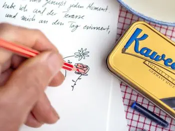 Jemand malt mit einem roten Stift eine gezeichnete Rose auf einem Brief ans Enkelkind aus. Daneben liegt eine goldene Blechdose von Kaweco. Foto: Johanna Rundel