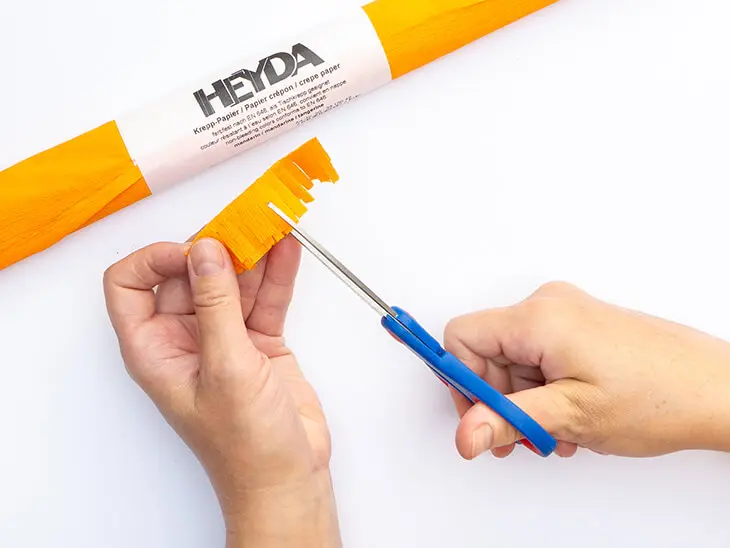 Jemand schneidet mit einer Schere Fransen in einen Streifen orangenes Krepppapier von HEYDA. Foto: Johanna Rundel