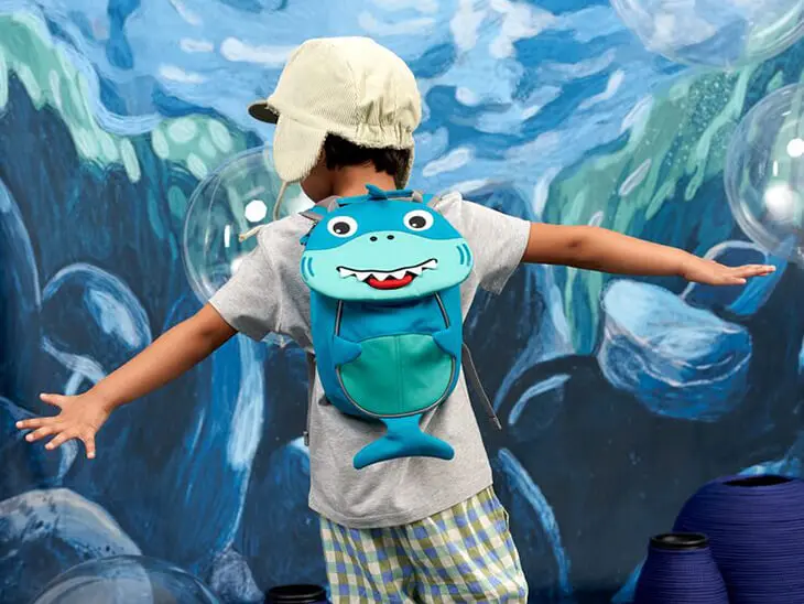 Ein Kleinkind tanzt mit seinem Rucksack in Haifischform von Affenzahn vor einer Unterwasserszene.