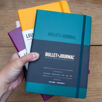 Jemand hält verschiedenfarbige Bullet Journals und Notizbücher von LEUCHTTURM1917 vor einem rustikalen Holzbrett in die Kamera. Foto: Johanna Rundel