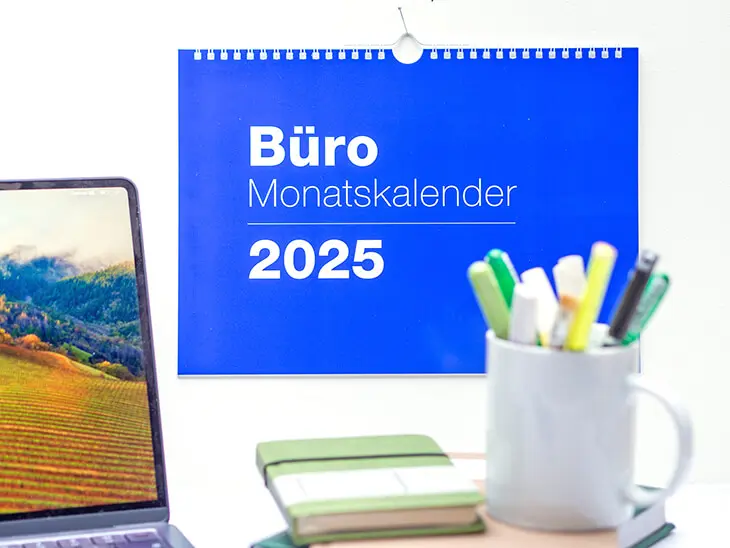 Ein blauer Büro-Monatskalender vom Athesia Kalenderverlag hängt an einer weißen Wand hinter einem Schreibtisch. Foto: Johanna Rundel