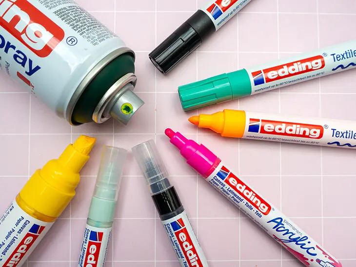 Verschiedene Marker, Stifte und Spraydosen von edding liegen im Kreis arrangiert auf einem rosa-weiß-karierten Untergrund. Foto: Johanna Rundel