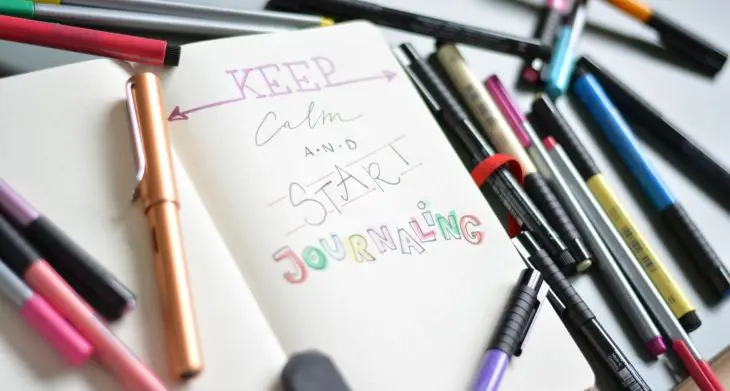Ein Bullet Journal liegt aufgeschlagen auf einem Tisch, umgeben von verschiedenen Stiften. Auf der Seite steht im Handlettering-Stil „Keep calm and start Journaling“.
