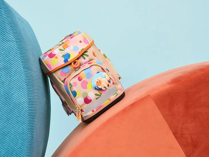 Ein rosa Schulranzen mit bunten Flecken von ergobag lehnt schräg zwischen zwei bunten Schaumstoffpolstern.) 