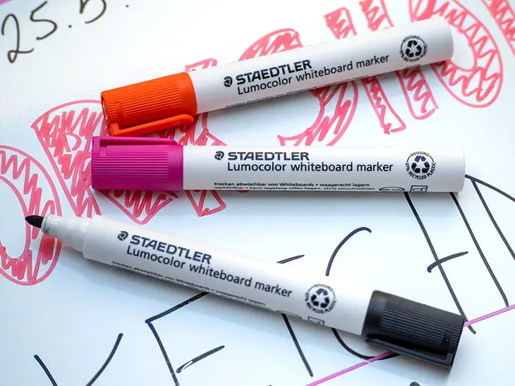 Ein roter, orangener und schwarzer Lumocolor Whiteboardmarker von STAEDTLER liegen auf einem beschrifteten Whiteboard. Foto: Johanna Rundel