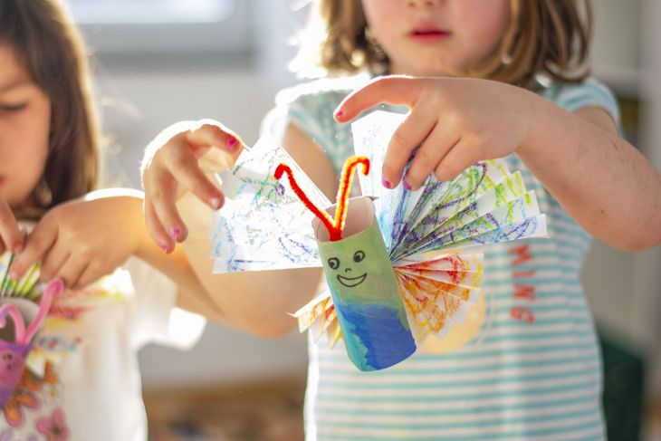 Zwei Kinder halten selbstgebastelte Schmetterlinge aus Klopapierrollen und buntem Papier in die Luft