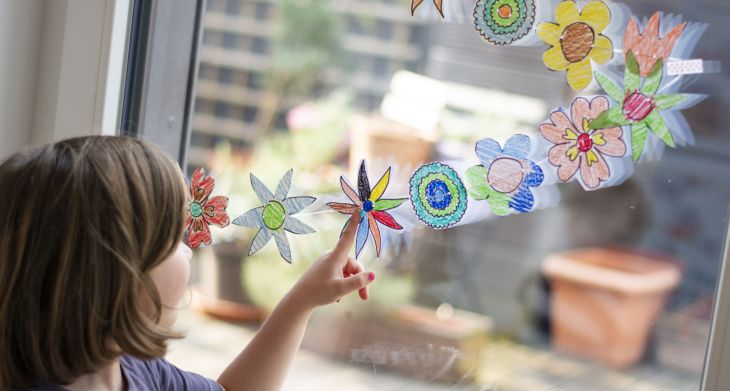 Ein Kind steht vor einem Fenster, auf das eine selbstgebastelte Blumengirlande geklebt ist und zeigt auf eine Blume