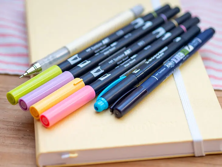 Ein hellgelbes Bullet Journal liegt auf einer Holzoberfläche. Auf dem Journal liegen verschiedene Stifte von Tombow. Es ist ein Starter-Set. Foto: Johanna Rundel