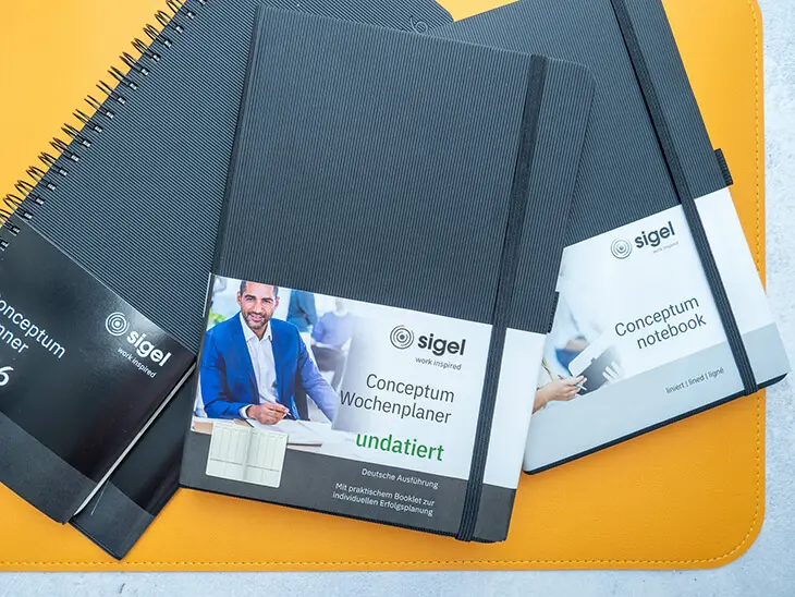 Ein Wochenplaner, ein undatierter Kalender und ein Notizbuch von Sigel liegen auf einer gelben Schreibtischunterlage. Foto: Johanna Rundel)