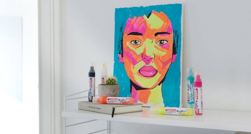 Bild eines Frauenkopfes, mit bunten Acrylmarkern gemalt, steht auf einer Kommode, darauf Dosen von edding Acrylmarkern.