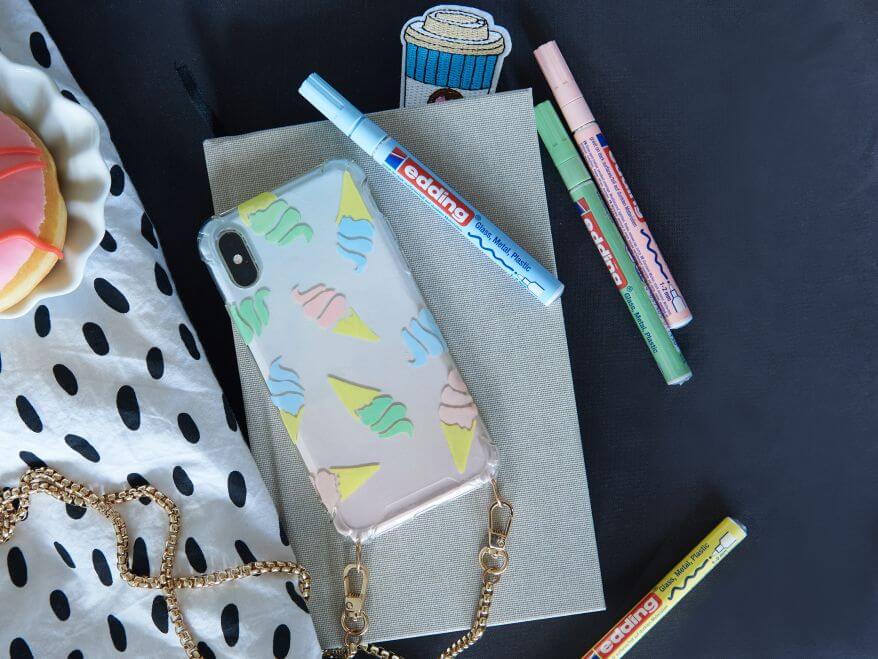 Durchsichtige Handyhülle wurde mit Glanzlackmarker von edding bemalt, welche neben dem Handy auf dem Tisch liegen.