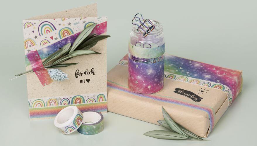 Eine selbstgebastelte Grußkarte und ein eingepacktea Geschenk stehen nebeneinander. Zentrale Gestaltungselemente sind verschiedene Dekotapes von Heyda in Regenbogenfarben. Die Karte ist mit dem Schriftzug "Für dich mit Herz" bestempelt, auf dem Geschenk steht "Überraschung". Dekoriert ist das Ensemble mit Salbeizweigen.