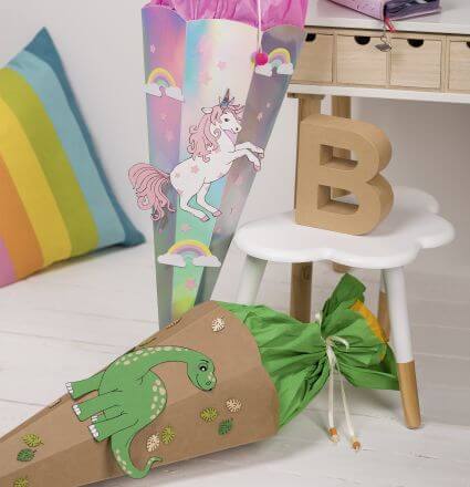 Man sieht zwei selbstgebastelte Schultüten aus Schultütensets der Marke Heyda. Auf dem Boden liegt eine braun-grüne Schultüte mit einem Dino, daneben steht eine Einhorn-Schultüte in Regenbogenfarben und rose. Drumherum sieht man etws Dekoration aus einem Kinderzimmer.
