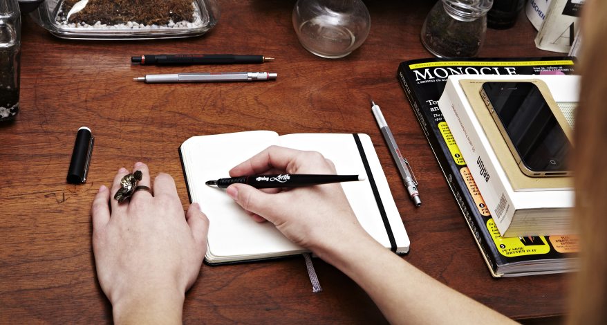 Jemnd schreibt mit einem Stift von Rotring in ein Notizbuch, welches auf einem Schreibtisch liegt.